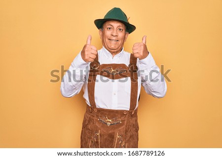 Senior grey-haired man wearing german traditional octoberfest suit over yellow background success sign doing positive gesture with hand, thumbs up smiling and happy. Cheerful expression and winner