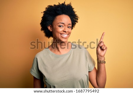 Young beautiful African American afro woman with curly hair wearing casual t-shirt with a big smile on face, pointing with hand and finger to the side looking at the camera.