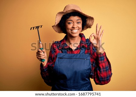 Young African American afro farmer woman with curly hair wearing apron and hat using rake doing ok sign with fingers, excellent symbol