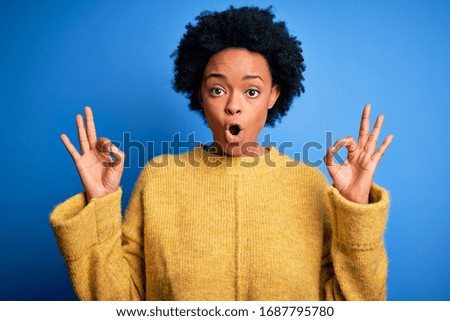 Young beautiful African American afro woman with curly hair wearing yellow casual sweater looking surprised and shocked doing ok approval symbol with fingers. Crazy expression