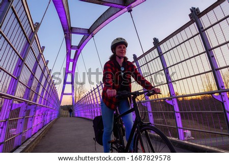 Caucasian Woman Riding a Bicycle on a Pedestrian Bridge over the Highway during a sunny sunset. Taken in Surrey, Vancouver, British Columbia, Canada. Alternative Transportation