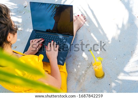 Girl in a yellow dress on a tropical sandy beach works on a laptop and drinks fresh mango. Remote work, successful freelance. Works on vacation