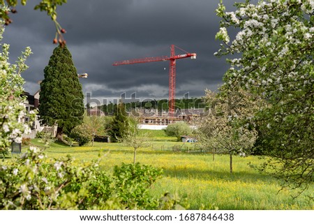 Red crane in stormy weather with sunlight. Illuminated green meadow with yellow spring flower and blooming orchard in sunlight. Dark stormy clouds and new residential district in background. Concept. 