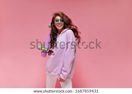 Attractive woman in white pants and purple hoodie looks into camera and smiles. Charming teen girl in green sunglasses holds colorful longboard.