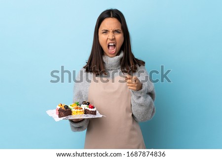 Pastry chef holding a big cake over isolated blue background frustrated and pointing to the front.