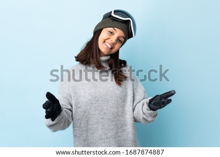 Mixed race skier girl with snowboarding glasses over isolated blue background presenting and inviting to come with hand.