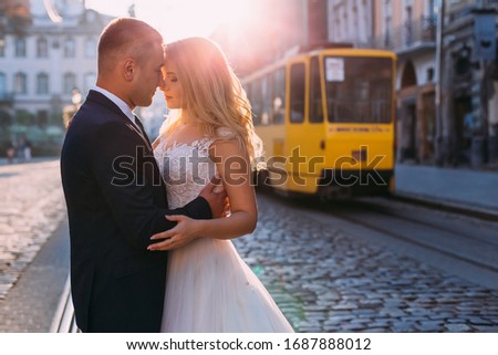 beautiful bride in white dress with lace. the groom in the suit hugs the bride. profile newlyweds on the background of square and tram.