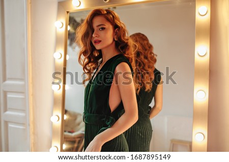 Cheerful woman in dark green outfit poses near mirror. Lovely lady with ginger wavy hair in stylish dress looking into camera.. Royalty-Free Stock Photo #1687895149