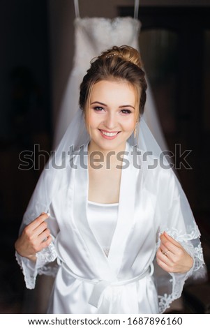 bride in white bathrobe with make up and bridal veil crossed arms and look at camera. wedding dress in the background. 