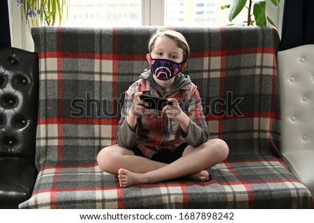 Photo of boy child in medical funny mask in quarantine in room at home plays on smartphone