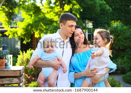 Happy young couple holding cute little kids while standing in yard of rural house with green trees in background in sunny summer day