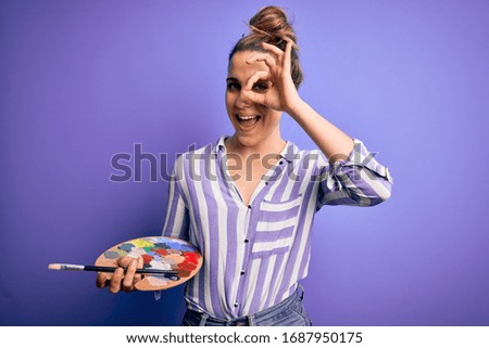 Young beautiful blonde artist woman painting using brush and palette over purple background with happy face smiling doing ok sign with hand on eye looking through fingers