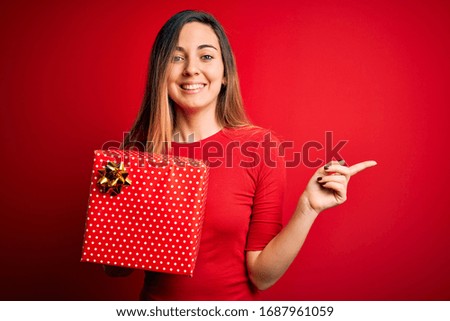 Young beautiful blonde woman with blue eyes holding birthday gift over red background very happy pointing with hand and finger to the side