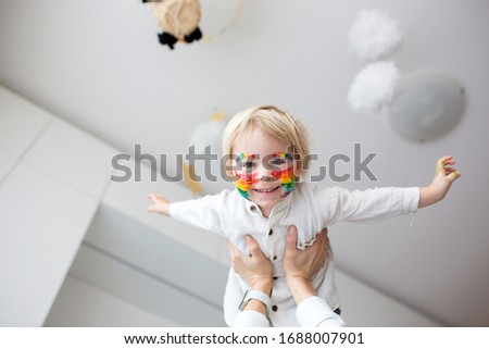 Beautiful blond toddler boy with rainbow painted on his face and messy hands, smiling happily