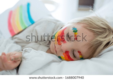 Beautiful blond toddler boy with rainbow painted on his face and messy hands, sleeping in his bed