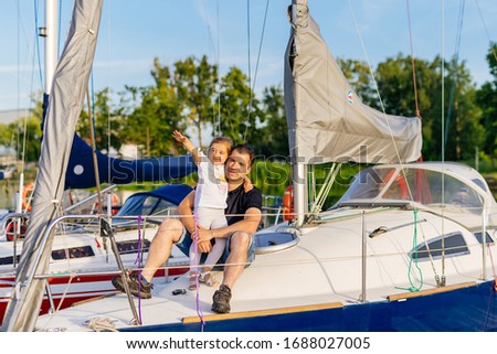 Portrait of smiling father and daughter on prow of sailboat or yacht anchored in marina at bright sunny day. Blurred background with boats and trees