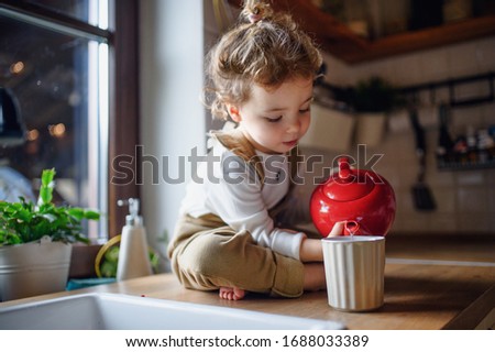 Cute small toddler girl sitting on kitchen counter indoors at home, pouring tea. Royalty-Free Stock Photo #1688033389