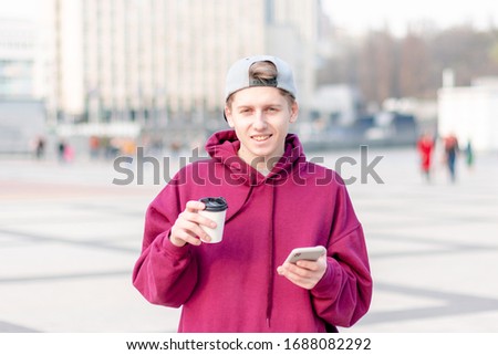 A large portrait of a young guy in a red sweater, in a good mood, holding coffee and a smartphone in his hands, looking at the camera. It stands against the backdrop of the square. 