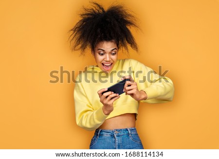 Overjoyed girl using smartphone, playing games. Photo of african american girl on yellow background