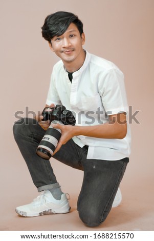 Asian man in white shirt kneeling while holding digital camera in hands. Male photographer looking at camera smiling. Isolated plain background copy space.