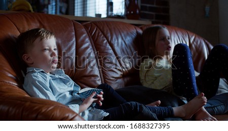 Entertainment and addiction, cute little Caucasian boy and girl watch TV at home on the couch with snacks slow motion.
