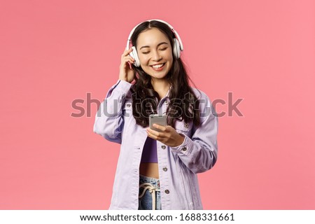 Portrait of stylish attractive and happy, smiling asian girl enjoying listening favourite songs in headphones, close eyes and laughing, found music station or funny podcast, hold mobile phone Royalty-Free Stock Photo #1688331661