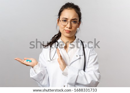 Portrait of displeased, serious-looking asian female doctor smirk skeptical, holding drugs, dont recommend taking pills or self-medicate, advice not use this medication, grey background Royalty-Free Stock Photo #1688331733