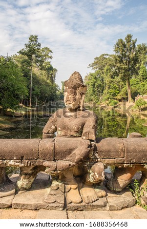 A large, ancient stone statue of a Buddhist god pulling on a giant snake forms the guardrail on a causeway leading to the famous Angkor Thom Temple in Siem Reap, Cambodia.
