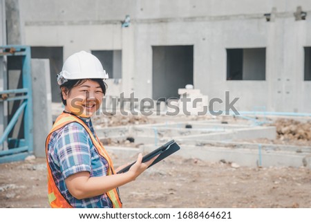 Asian woman civil construction engineer worker or architect with helmet and safety vest working and holding a touchless tablet computer for see blueprints or plan at a building or construction site