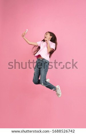 Jumping high, selfie. Caucasian little girl portrait isolated on pink studio background. Cute brunette model in shirt. Concept of human emotions, facial expression, sales, ad, childhood. Copyspace.