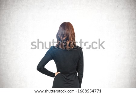 Rear view of young businesswoman in black dress with wavy brown hair looking at concrete wall. Concept of advertising