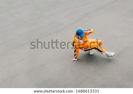 Photo of stylish street dancer showing breakdancing performance on asphalt background. Young man in orange clothes dancing hip hop on the street. Isolated on gray asphalt background