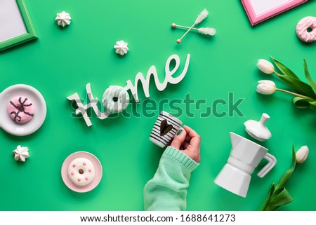 Stay home message, creative flat lay on green mint background with Spring objects and symbols. Hand holding espresso cup with heart sign. Word "Home", coffee maker, cups and white marshmallow sweets