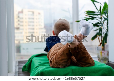 Child boy lies on a blanket on the balcony and looks out the window. Isolation from the outside world. World Quarantine, Coronovirus Pantemia, COVID-19. To stay home. Preventive measures. Danger
