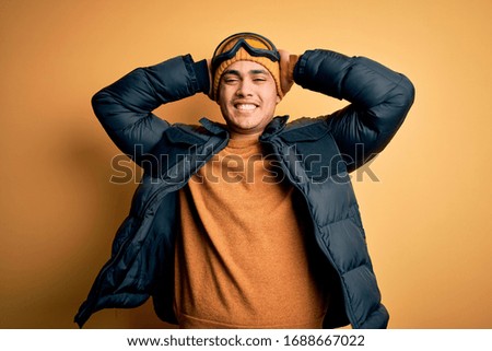 Young brazilian skier man wearing snow sportswear and ski goggles over yellow background relaxing and stretching, arms and hands behind head and neck smiling happy