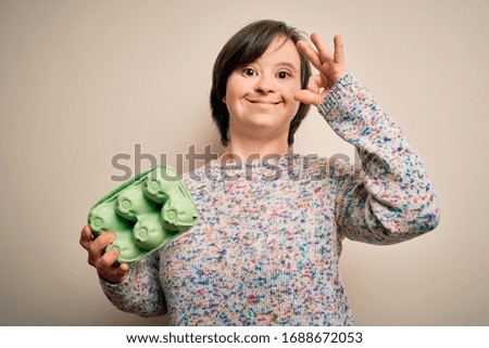 Young down syndrome woman holding cardboard egg cup from fresh healthy eggs doing ok sign with fingers, excellent symbol