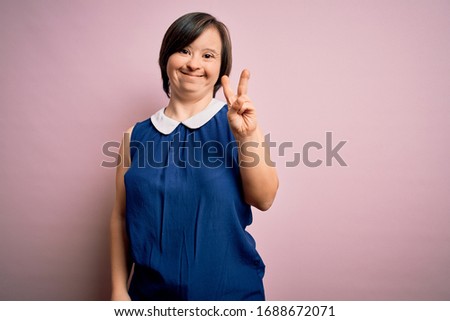 Young down syndrome woman wearing elegant shirt over pink background smiling with happy face winking at the camera doing victory sign. Number two.