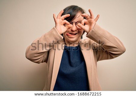 Young down syndrome business woman wearing glasses standing over isolated background doing ok gesture like binoculars sticking tongue out, eyes looking through fingers. Crazy expression.