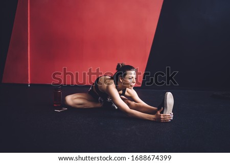 Focused fit female in dark sporty top and short shorts stretching muscles while bending forward to one leg sitting on floor in gym looking away