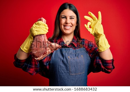Young butcher woman holding fresh raw beef meat stake over red background doing ok sign with fingers, excellent symbol