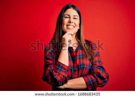 Young beautiful woman wearing casual shirt over red background looking confident at the camera with smile with crossed arms and hand raised on chin. Thinking positive.
