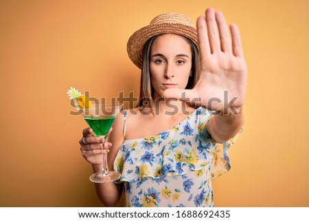 Young beautiful tourist woman on vacation wearing summer hat drinking cocktail beverage with open hand doing stop sign with serious and confident expression, defense gesture