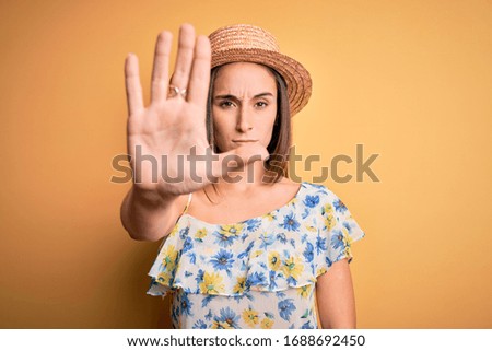 Young beautiful woman wearing casual t-shirt and summer hat over isolated yellow background doing stop sing with palm of the hand. Warning expression with negative and serious gesture on the face.