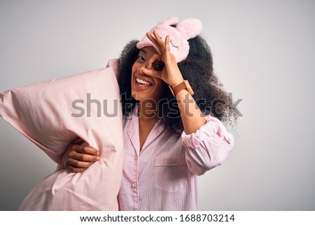 Young african american woman with afro hair wearing pajama and sleep mask holding pillow with happy face smiling doing ok sign with hand on eye looking through fingers