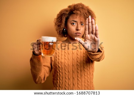 Young beautiful African American afro woman with curly hair drinking jar of beer with open hand doing stop sign with serious and confident expression, defense gesture