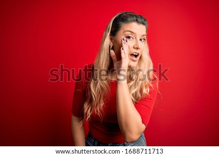 Young beautiful blonde woman wearing casual t-shirt standing over isolated red background hand on mouth telling secret rumor, whispering malicious talk conversation