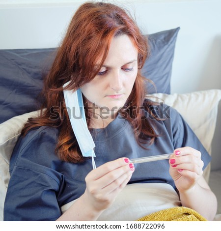Redhead woman with coronavirus with mask on her face looks at a thermometer. Girl with a flu virus in bed at home in a medical mask