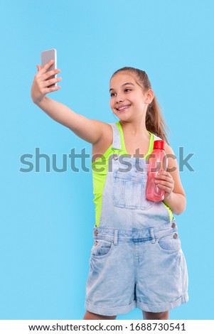 Shot of young brunette girl wearing  denim overalls shorts standing with a phone and with a bottle of water. She takes a selfie. Vacation and reacriation concept
