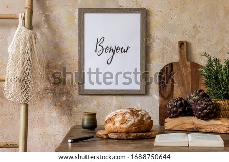 Stylish interior of kitchen space with wooden table, brown mock up photo frame, bread, cup of tea, basket, book, vegetable and kitchen accessories in wabi sabi concept of home decor. 