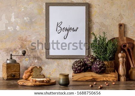 Stylish interior of kitchen space with wooden table, brown mock up photo frame, bread, cup of tea, herbs, vegetables and kitchen accessories in wabi sabi concept of home decor. 